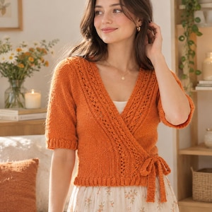 May include: A burnt orange knit wrap top featuring short sleeves, a V-neckline, and a side tie closure. The garment is constructed with a combination of stockinette stitch and vertical lace detailing along the front edges and hemline.