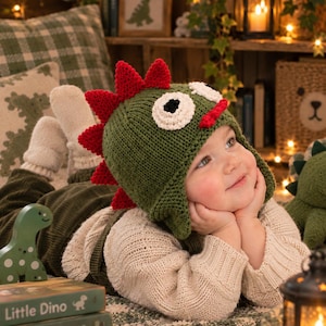 May include: A green knitted hat featuring a dinosaur design with red spikes along the top and back, large white and black circular eyes, and ear flaps. The hat is crafted with a textured knit pattern and includes a small red mouth detail on the front.