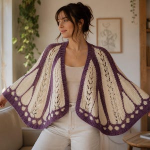 May include: A knitted open-front cardigan featuring a cream and purple butterfly wing pattern. The garment has a wide, draped silhouette with intricate lace-like stitch details in the cream sections and solid purple borders along the edges.