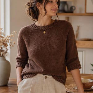 Könnte beinhalten: Ein dunkelbrauner Strickpullover mit Rundhalsausschnitt, Raglanärmeln sowie gerippten Bündchen an Ärmeln und Saum. Der Pullover wird mit einer hellbeigen Hose kombiniert, die einen sichtbaren Knopfverschluss an der Taille aufweist.