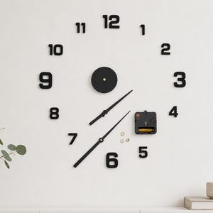 Op de afbeelding: Een wandklokset met individuele zwarte zelfklevende cijfers van 1 tot 12, een centrale ronde naaf, twee zwarte wijzers en een uurwerk op batterijen met drie kleine metalen ringetjes voor de montage en installatie van de klok.
