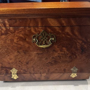 May include: A rectangular wooden box with a rich, swirling grain pattern in shades of brown. The box features a prominent, ornate brass handle centered on the front panel and two small, decorative brass hinges at the bottom edge of the lid.