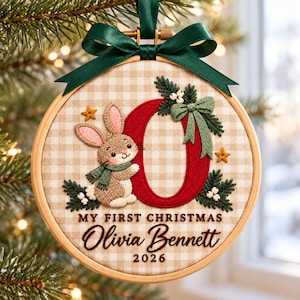 May include: A round wooden embroidery hoop ornament featuring a beige and white gingham fabric background. It displays a brown bunny, a large red letter O, green pine sprigs, and white berries. Text reads: MY FIRST CHRISTMAS Olivia Bennett 2026.