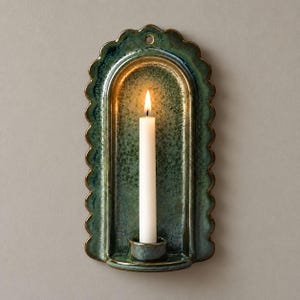 May include: A wall-mounted ceramic candle sconce with a scalloped edge and a glossy, mottled green glaze. The arched backplate features a central hole for hanging and a small circular base at the bottom holding a single, lit white taper candle.