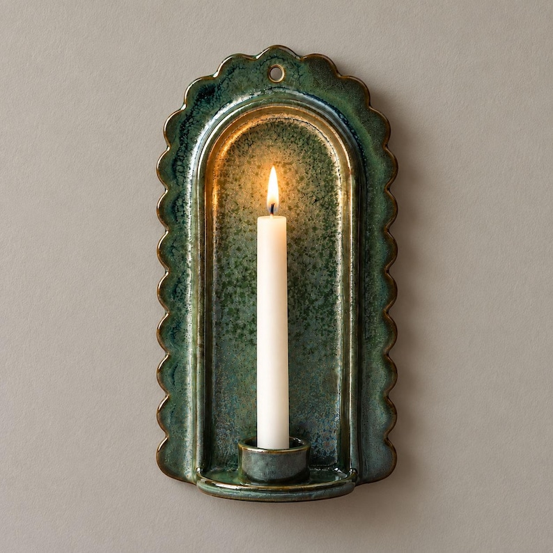 May include: A wall-mounted ceramic candle sconce with a scalloped edge and a glossy, mottled green glaze. The arched backplate features a central hole for hanging and a small circular base at the bottom holding a single, lit white taper candle.