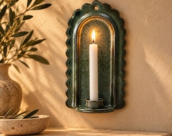 May include: A wall-mounted ceramic candle holder with a scalloped edge and a glossy, mottled dark green glaze. The arched backplate features a small hole at the top for hanging, and a central circular base holds a single, slender white taper candle.