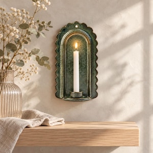 May include: A wall-mounted arched candle holder with a scalloped edge and a mottled dark green glaze. The holder features a central circular cup containing a single white taper candle with a lit flame, and a small hole at the top for mounting.