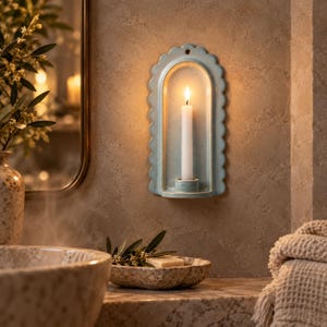 May include: A light blue ceramic wall sconce with a scalloped edge and an arched frame. The sconce features a small circular hole at the top for mounting and a central cylindrical holder containing a single white taper candle with a lit flame.