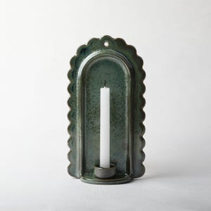 May include: A wall-mounted ceramic candle holder with a scalloped edge and a deep green, mottled glaze. The arched backplate features a small hole at the top for hanging, and a cylindrical base holds a single, slender white taper candle.
