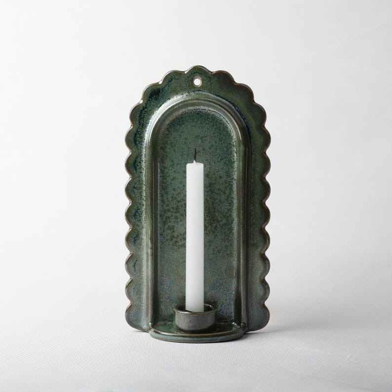 May include: A wall-mounted ceramic candle holder with a scalloped edge and a deep green, mottled glaze. The arched backplate features a small hole at the top for hanging, and a cylindrical base holds a single, slender white taper candle.