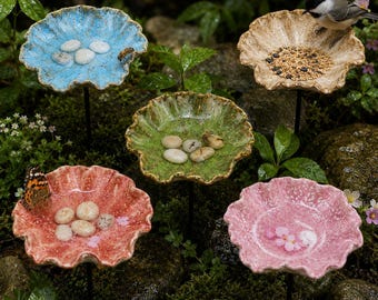 May include: Five circular, scalloped-edge garden dishes on thin metal stakes. The dishes are glazed in blue, beige, green, red, and pink, featuring textured surfaces. Some contain small white stones, birdseed, or a butterfly and bee.