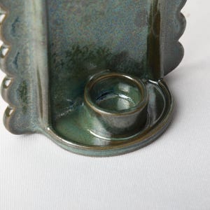 May include: A dark teal ceramic candle holder featuring a glossy, mottled glaze finish. The object has a central circular cup for a candle, surrounded by a curved back panel with scalloped, wavy edges on both sides, creating a distinct silhouette.