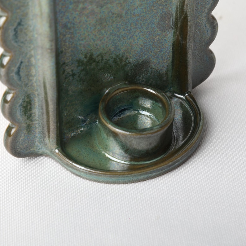 May include: A dark teal ceramic candle holder featuring a glossy, mottled glaze finish. The object has a central circular cup for a candle, surrounded by a curved back panel with scalloped, wavy edges on both sides, creating a distinct silhouette.