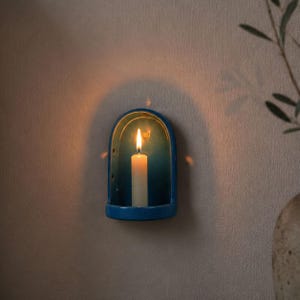 May include: A wall-mounted arched candle holder in a deep blue glaze. The holder features a curved, semi-circular frame with a flat base, containing a single off-white pillar candle with a lit flame that casts a warm glow against the wall.