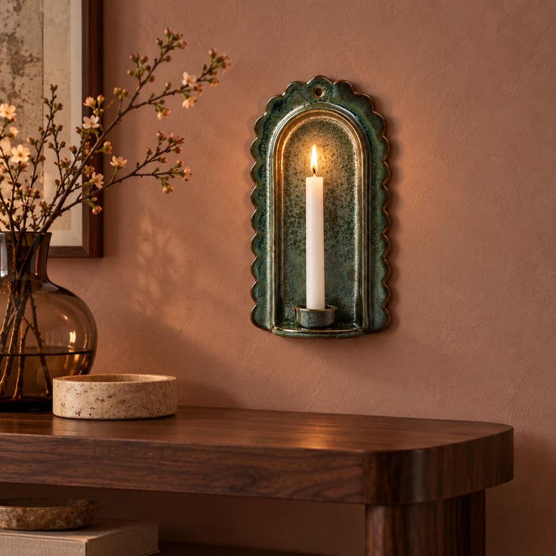 May include: A wall-mounted ceramic candle sconce with a scalloped edge and a deep green, mottled glaze. The arched piece features a small circular hole at the top for mounting and a central platform holding a single, lit white taper candle.