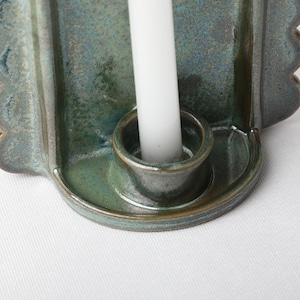 May include: A ceramic candle holder with a glossy, mottled blue-green glaze featuring a scalloped edge design. A single, plain white taper candle is inserted into the central circular socket of the holder, which has a smooth, curved base.