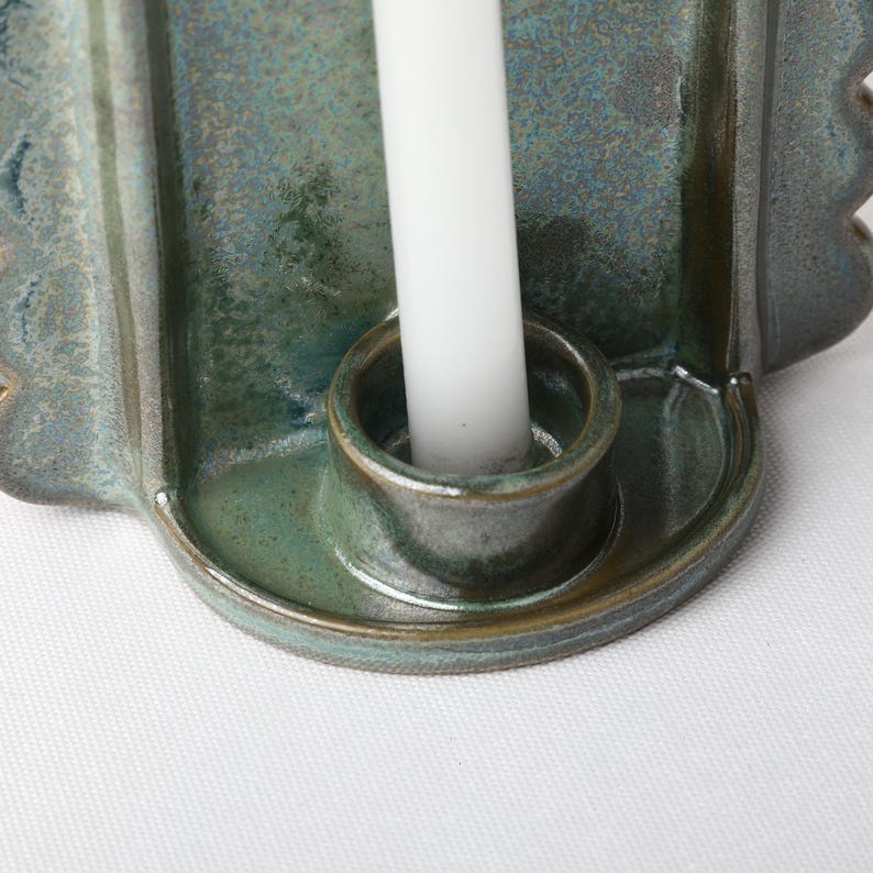 May include: A ceramic candle holder with a glossy, mottled blue-green glaze featuring a scalloped edge design. A single, plain white taper candle is inserted into the central circular socket of the holder, which has a smooth, curved base.