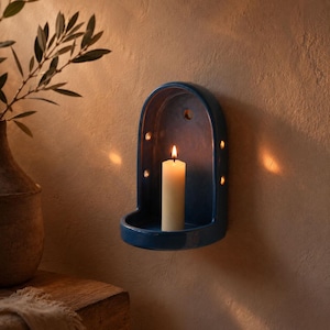 May include: A deep blue ceramic wall-mounted candle holder with an arched top and a flat base. The holder features small circular cutouts along the sides and a single hole at the top for mounting. A lit white pillar candle sits inside the holder.