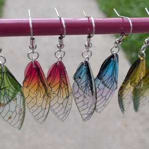 May include: Eight dangling earrings shaped like insect wings, featuring translucent resin with intricate black vein patterns and gold fleck accents. Each earring hangs from a silver-toned metal hook, displaying a gradient of green, pink, orange, blue, and yellow hues.