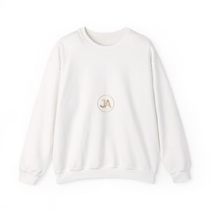 May include: A long-sleeve crewneck sweatshirt in a solid off-white color. Centered on the chest is a circular emblem featuring the gold-colored letters JA inside a thin gold-colored ring. The garment has ribbed cuffs and a ribbed hemline.