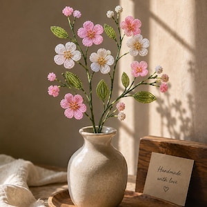 以下が含まれることがあります： 小さな明るい色の陶器の花瓶に飾られた、ビーズで作られた花の装飾品。花は黄色い中心部を持つピンクと白の花びらで構成され、細い茎には緑色のビーズの葉が付いています。小さなカードには「Handmade with love」と書かれています。