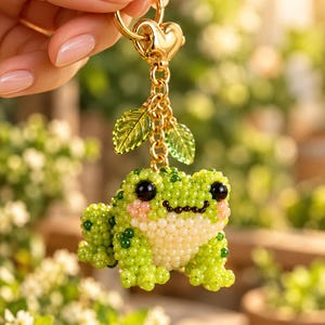 Könnte beinhalten: Ein Perlen-Schlüsselanhänger in Form eines kleinen Frosches, der aus winzigen grünen und cremefarbenen Perlen gefertigt ist. Der Frosch hat große schwarze Augen und rosa Wangen. Er hängt an einer goldfarbenen Metallkette mit zwei durchsichtigen grünen Blattanhängern.