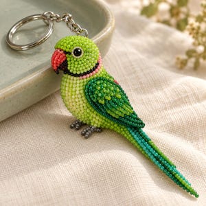 May include: A beaded parrot keychain featuring a vibrant green body, a red beak, and a long tail made of small, uniform beads. The bird has a black eye and a silver-toned metal keyring attached to the top by a short chain link.