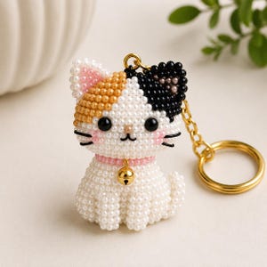 Può includere: Un portachiavi a forma di gatto calico realizzato con piccole perline bianche, arancioni e nere. Il gatto presenta occhi di perline nere, guance di perline rosa e un collare di perline rosa con un piccolo campanello in metallo dorato.