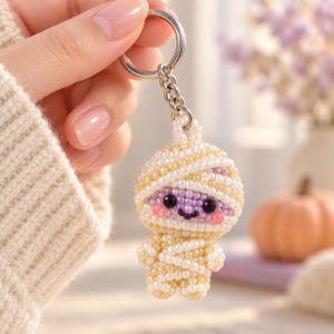 May include: A keychain featuring a small, handmade figure crafted from cream-colored beads wrapped to resemble a mummy. The figure has a light purple face, two black bead eyes, and a small dark purple stitched smile with pink bead cheeks.
