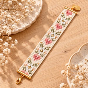 Könnte beinhalten: Ein breites Perlenarmband mit einem sich wiederholenden Muster aus rosa Herzen und weißen Blumen mit grünen Blättern auf cremefarbenem Grund. Das Armband ist mit goldfarbenen Metallendstücken und einem kleinen runden Anhänger versehen.