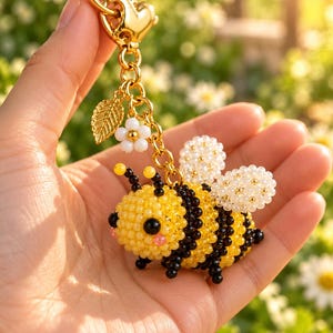 Puede incluir: Un llavero con forma de abeja hecho de cuentas amarillas, negras y blancas. La abeja tiene una cabeza amarilla con ojos negros, un cuerpo a rayas amarillas y negras y alas de cuentas blancas. Cuelga de una cadena de metal dorado con un dije de hoja.