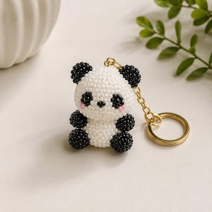 May include: A panda keychain crafted from small white and black beads. The panda figure features a round white head with black ears, black eyes, a small black nose, and pink accents on the cheeks. It has black limbs and a gold-toned metal ring.