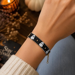 Könnte beinhalten: Ein Perlenarmband mit schwarzem Hintergrund und einem sich wiederholenden Muster aus weißen Geistern und kleinen goldfarbenen Kreuzen. Das Armband verfügt über eine goldfarbene Metallkettenverlängerung mit Karabinerverschluss.
