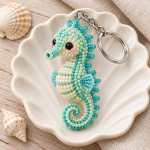 May include: A seahorse keychain crafted from small, iridescent beads in shades of mint green, turquoise, and cream. The seahorse features two black bead eyes and small pink bead accents on its cheeks, attached to a silver metal keyring.