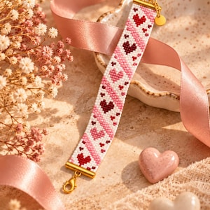 May include: A woven bead bracelet featuring a repeating pattern of red and pink hearts on a white background. The bracelet is finished with gold-toned metal end caps, a lobster claw clasp, and a small circular charm attached to one end.