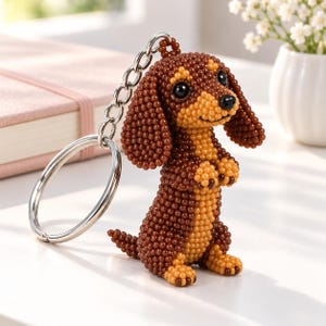 Op de afbeelding: Een teckel-sleutelhanger gemaakt van kleine bruine en beige kralen. Het hondje zit rechtop met de voorpootjes omhoog, heeft oogjes en een neus van zwarte kralen en is bevestigd aan een zilverkleurige metalen ring.
