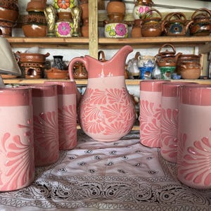 May include: A ceramic pitcher and six matching tumblers in a soft pink hue. Each piece features a raised, embossed floral pattern in a lighter shade of pink. The pitcher has a curved handle and a spout, while the tumblers are cylindrical.