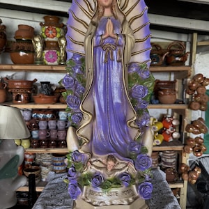 Pode incluir: Uma estátua alta e vertical da Virgem de Guadalupe com um manto roxo, capa cor de creme e uma auréola dourada em forma de sol. A figura é rodeada por rosas roxas e posicionada sobre um pequeno anjo, com o texto BENDICE ESTE HOGAR.