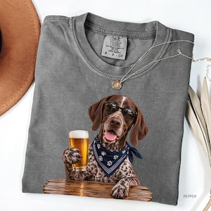 May include: A charcoal gray crew neck t-shirt featuring a graphic of a brown and white dog wearing sunglasses and a blue bandana with white paw prints. The dog is holding a glass of beer while resting its paws on a wooden surface. The shirt has a Comfort Colors label.