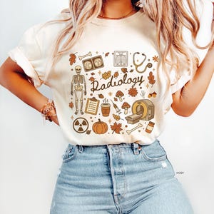May include: An ivory t-shirt featuring a brown graphic print with the word Radiology in cursive script. The design includes a skeleton, medical equipment like a stethoscope, syringe, and CT scanner, alongside autumn elements like leaves, acorns, and a pumpkin.