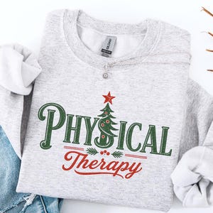 May include: A light gray crewneck sweatshirt featuring the text Physical Therapy in dark green and red. The letter S is replaced by a green Christmas tree with a red star on top. The word Therapy is in red script below the tree.
