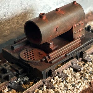May include: A weathered model train flatcar featuring a cylindrical brown tank with visible rust textures, a circular perforated metal plate, and horizontal rod details. The miniature railway car is positioned on a set of tracks with gravel ballast.