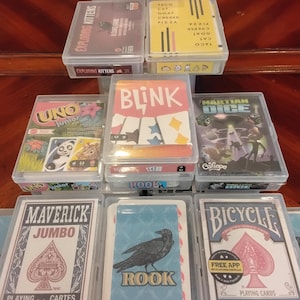 May include: A collection of various card games and playing cards stored in clear plastic cases, including Exploding Kittens, Taco Cat Goat Cheese Pizza, Uno Junior, Blink, Martian Dice, Maverick Jumbo, Rook, and Bicycle Standard playing cards.