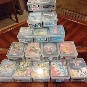 May include: A pyramid-shaped stack of clear plastic storage cases containing various card games, including Five Crowns, Mille Bornes, Sea Salt & Paper, Waterworks, Flip 7, CuBirds, Ruckus, Skull King, Bears vs Bees, Dragonwood, Cover Your Assets, Uno Flex, Uno Teams, Uno H2O To Go, and Phase 10.