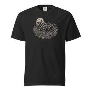 May include: A black short-sleeve t-shirt featuring a cream-colored graphic print of a sea otter floating in water, surrounded by a wreath of pine needles and pinecones. The design is centered on the chest area of the garment.