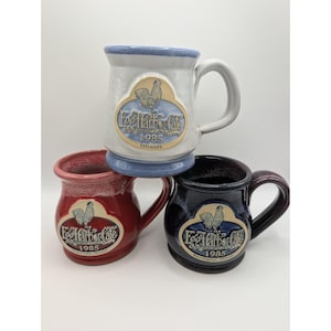 May include: Three ceramic mugs in white, red, and dark blue, each featuring a raised emblem with a rooster illustration and the text Egg Harbor Cafe, Breakfast & Lunch, 1985, and the location name Illinois, Naperville, or Barrington.