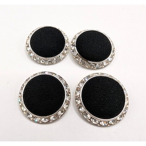 May include: Four circular buttons featuring a black fabric center surrounded by a silver-toned metal rim embedded with clear, faceted crystals. The buttons are arranged in a two-by-two grid, showcasing a consistent design with a textured black surface.