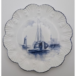 May include: A white ceramic plate with a scalloped edge and a dark blue rim. The center features a blue ink illustration of a sailboat on water with a distant shoreline and a tower. The plate border has a raised, embossed fan-like pattern.