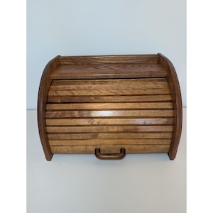 May include: A medium-brown wooden bread box featuring a curved roll-top door with horizontal slats and a small matching handle. The unit includes a flat top shelf and a sturdy frame, showcasing a natural wood grain finish throughout the structure.