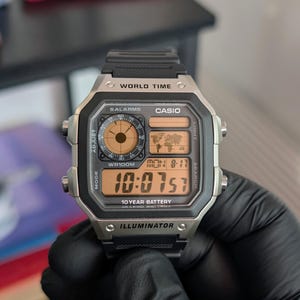 Op de afbeelding: Een digitaal Casio-horloge met een zilverkleurige rechthoekige kast en een zwarte kunsthars band. De wijzerplaat bevat een wereldkaart, een ronde analoge sub-wijzerplaat en een digitale tijdweergave van 10:07:57 op een amberkleurige achtergrond.